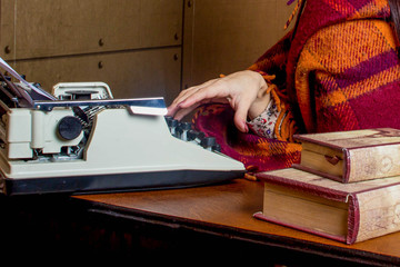 Girl typing on a typewriter. On the table are also two books. Girl wrapped in a plaid
