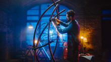 Handsome Male Artist Uses An Angle Grinder To Make Brutal Metal Sculpture In Studio. Hipster Guy Polishes Metal Tube With Sparks Flying Off It. Contemporary Fabricator Creating Abstract Steel Art.