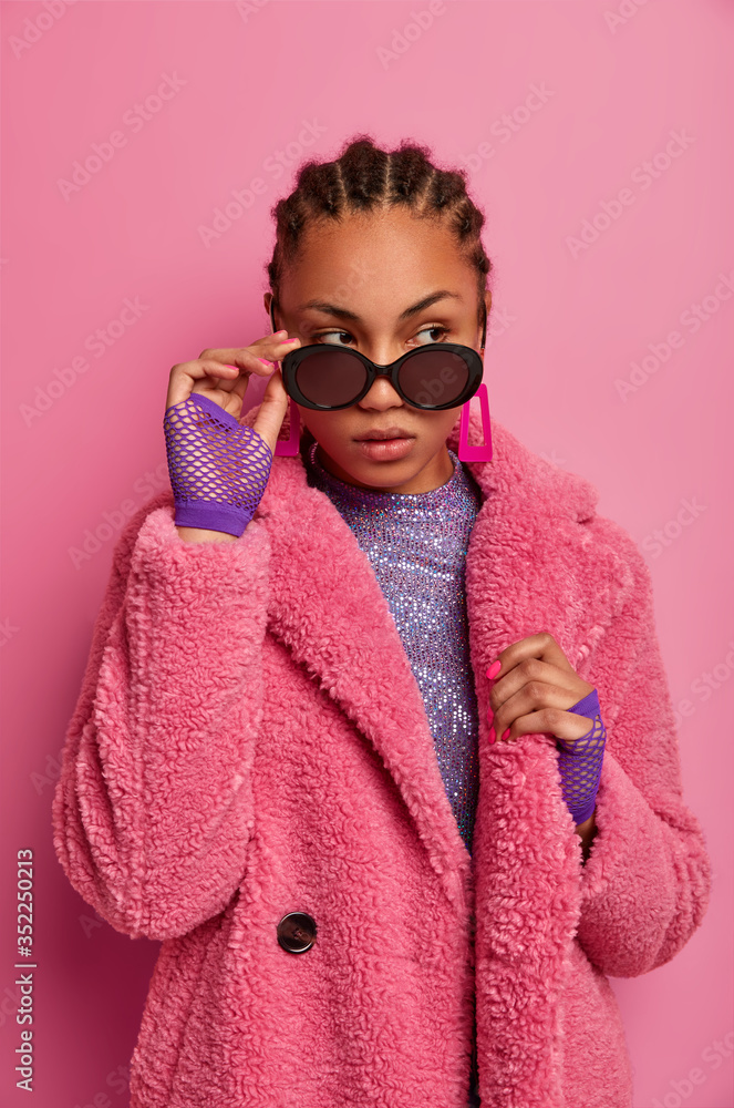 Vertical shot of thoughtful dark skinned female fashion model being well dressed, wears stylish coat and sunglasses, looks pensively aside with confident expression, isolated over rosy background - obrazy, fototapety, plakaty 