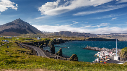 Leinwandbilder - Amazing nature of Iceland. View of the basalt rocks formation on the coastline with beautiful houses and the Mountain Stapafell in the background at Arnarstapi Village in summer sunny day in Iceland