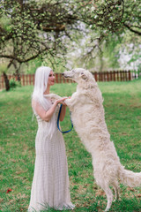 Blonde woman in white dress playing with her russian wolfhound dog in garden.