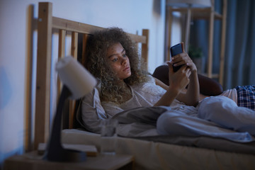 Wall Mural - Portrait of young curly-haired woman using smartphone in bed at night while suffering from insomnia, copy space