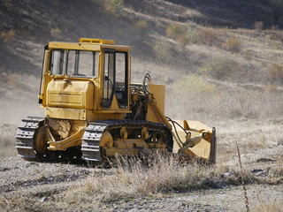 A large yellow bulldozer flattens a mountain dirt road in clouds of dust on a Sunny autumn day