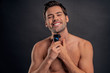 © Roman - Handsome young bearded man isolated. Portrait of topless muscular man is standing on gray background with electric Shaver in hand while shaving. Men care concept.