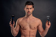 Handsome young bearded man isolated. Portrait of topless muscular man is standing on gray background with trimmer in one hand and razor in other in hand. Men care concept.