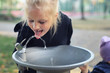 Cute adorable caucasian blond little thirsty school girl drinking water from public potable fountain faucet in city park on bright hot summer day outdoors. Children dehydration at heat season