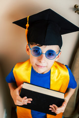 little cute preschooler boy in glasses with books at home education in graduate hat smiling posing emotional, lifestyle people concept