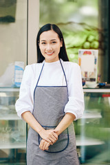 Wall Mural - Portrait of young lovely Asian barista in apron standing in front of coffeeshop