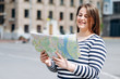 Young joyful woman with tourist map in hands enjoying beautiful strolling during her spring journey, happy female with cute smile studying atlas before walking in foreign city during summer trip