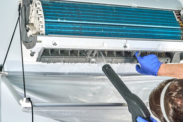 Engineer in blue gloves and mask cleaning Gree dirty air conditioner from dust and mold with water vacuum cleaner.