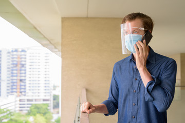 Poster - Young man talking on the phone with mask and face shield against view of the city