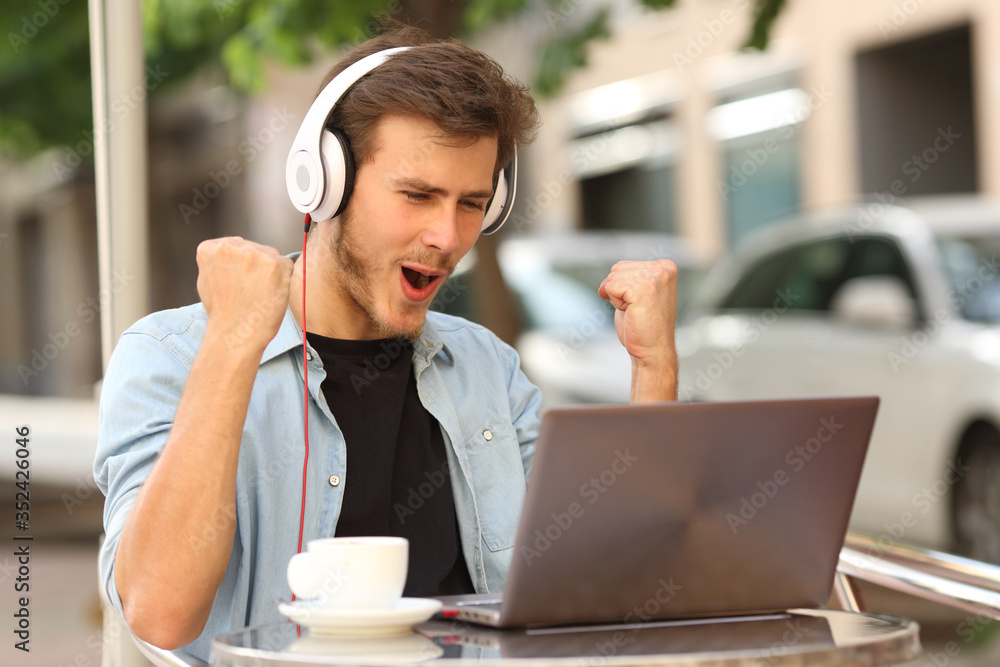 Man with headphones celebrating news on laptop on a bar - obrazy, fototapety, plakaty 