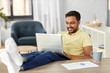 © Syda Productions - technology, remote job and lifestyle concept - happy smiling indian man with laptop computer resting feet on table at home office