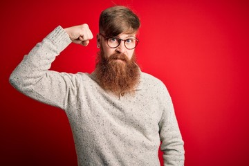 Canvas Print - Handsome Irish redhead man with beard wearing casual sweater and glasses over red background Strong person showing arm muscle, confident and proud of power