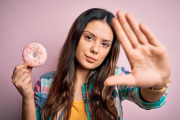 Wall Mural - Young beautiful brunette woman eating sweet pink doughnut over isolated background with open hand doing stop sign with serious and confident expression, defense gesture