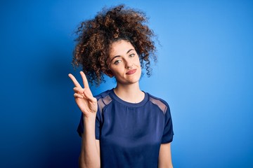 Young beautiful woman with curly hair and piercing wearing casual blue t-shirt showing and pointing up with fingers number two while smiling confident and happy.