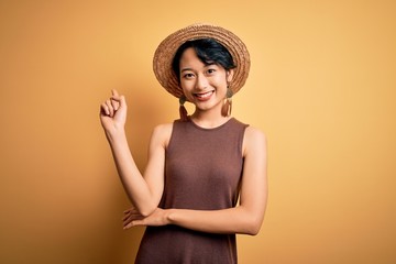 Poster - Young beautiful asian girl wearing casual t-shirt and hat over isolated yellow background with a big smile on face, pointing with hand and finger to the side looking at the camera.