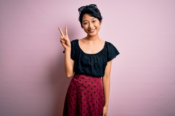 Wall Mural - Young beautiful asian girl wearing casual dress standing over isolated pink background smiling with happy face winking at the camera doing victory sign with fingers. Number two.