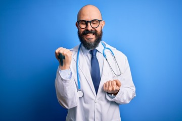 Wall Mural - Handsome bald doctor man with beard wearing glasses and stethoscope over blue background celebrating surprised and amazed for success with arms raised and eyes closed. Winner concept.