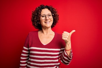 Poster - Middle age beautiful curly hair woman wearing casual striped sweater over red background smiling with happy face looking and pointing to the side with thumb up.