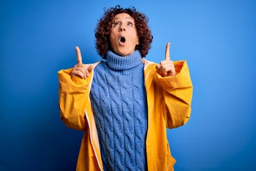 Poster - Middle age curly hair woman wearing rain coat standing over isolated blue background amazed and surprised looking up and pointing with fingers and raised arms.