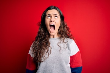 Sticker - Young beautiful woman with curly hair wearing casual sweatshirt over isolated red background angry and mad screaming frustrated and furious, shouting with anger. Rage and aggressive concept.