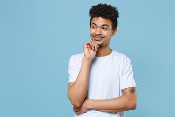 Smiling young african american guy in casual white t-shirt posing isolated on blue background studio portrait. People lifestyle concept. Mock up copy space. Put hand prop up on chin, looking aside.