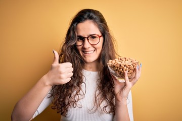 Sticker - Beautiful woman with curly hair holding bowl with healthy peanuts over yellow background happy with big smile doing ok sign, thumb up with fingers, excellent sign