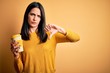 © Krakenimages.com - Young woman with blue eyes holding cup of coffee standing over yellow background with angry face, negative sign showing dislike with thumbs down, rejection concept