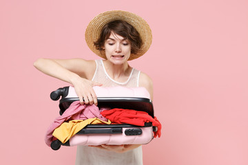 Confused young tourist girl in summer white dress hat isolated on pink background studio portrait. Female traveling abroad to travel weekend getaway. Air flight journey concept. Hold opened suitcase.