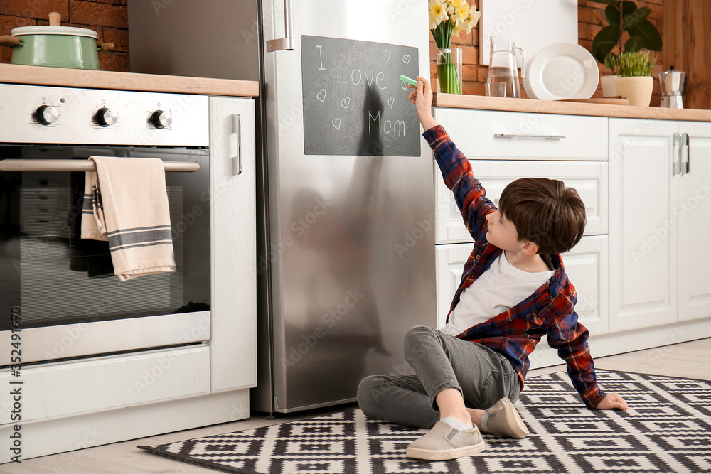 Little boy writing text I LOVE MOM on chalkboard in kitchen