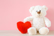 Smiling white teddy bear sitting and holding red soft heart at pastel pink wall. Front view. Closeup.
