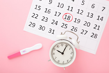 Canvas Print - Calendar, test with two stripes and white alarm clock on light pink table background. Pastel color. Positive result. Pregnancy time beginning. Closeup. Top down view.