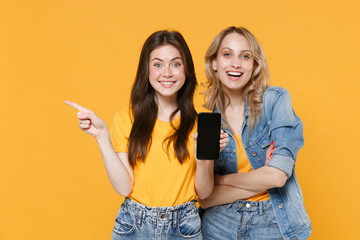 Two smiling young women girls friends in casual t-shirts denim clothes isolated on yellow background. People lifestyle concept. Hold mobile phone with blank empty screen, pointing index finger aside.