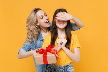 Funny young women girls friends in casual t-shirts denim clothes isolated on yellow background. Women's Day birthday, holiday concept. Hold present box with gift ribbon bow, covering eyes with hand.