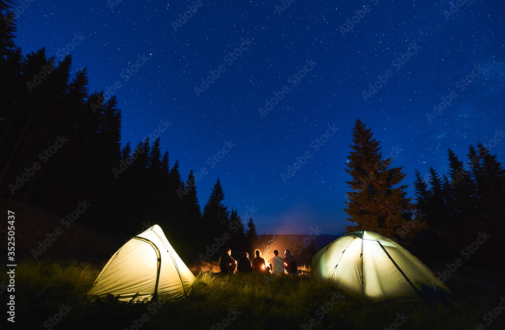 Back view, tourists sitting by brightly blazing campfire between tents against the backdrop of mountain valley and forest under blue night sky with bright stars. Concept of tourism, evening camping - obrazy, fototapety, plakaty 