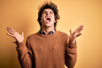 Wall Mural - Young handsome man wearing casual shirt and sweater over isolated yellow background crazy and mad shouting and yelling with aggressive expression and arms raised. Frustration concept.