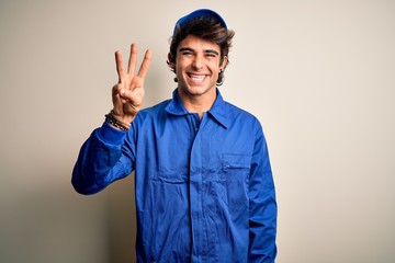 Wall Mural - Young mechanic man wearing blue cap and uniform standing over isolated white background showing and pointing up with fingers number three while smiling confident and happy.