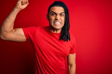 Wall Mural - Young handsome african american afro man with dreadlocks wearing red casual t-shirt angry and mad raising fist frustrated and furious while shouting with anger. Rage and aggressive concept.