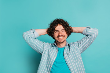 Relaxed positive cheerful young man isolted over blue background. Handsome guy hold hands behind head and smiling on camera. Posing alone in studio.