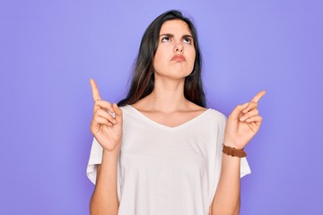 Poster - Young beautiful brunette woman wearing casual white t-shirt over purple background Pointing up looking sad and upset, indicating direction with fingers, unhappy and depressed.