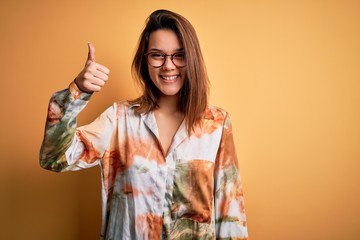 Young beautiful brunette girl wearing casual shirt and glasses over isolated yellow background doing happy thumbs up gesture with hand. Approving expression looking at the camera showing success.