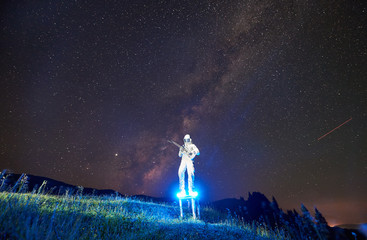 Wall Mural - Astronaut with glowing boots playing guitar under starry sky with Milky Way. Cosmonaut guitarist with musical instrument wearing white space suit and helmet. Concept of music, cosmonautics and galaxy.