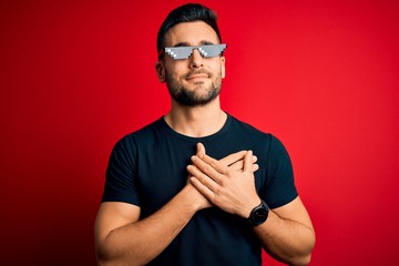 Sticker - Young handsome man wearing funny thug life sunglasses over isolated red background smiling with hands on chest with closed eyes and grateful gesture on face. Health concept.