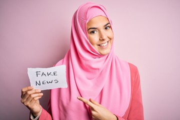 Poster - Young beautiful girl wearing muslim hijab holding paper with fake news message very happy pointing with hand and finger