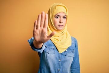 Wall Mural - Young beautiful girl wearing muslim hijab standing over isolated yellow background doing stop sing with palm of the hand. Warning expression with negative and serious gesture on the face.