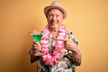 Grey haired senior man wearing summer hat and hawaiian lei drinking a cocktail very happy pointing with hand and finger