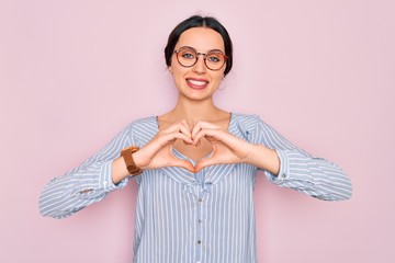 Wall Mural - Young beautiful woman wearing casual striped shirt and glasses over pink background smiling in love doing heart symbol shape with hands. Romantic concept.