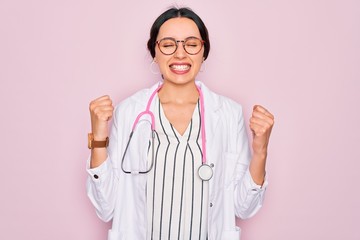 Wall Mural - Beautiful doctor woman with blue eyes wearing coat and stethoscope over pink background very happy and excited doing winner gesture with arms raised, smiling and screaming for success. Celebration