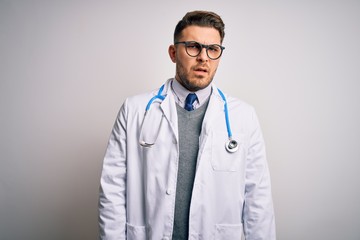 Wall Mural - Young doctor man with blue eyes wearing medical coat and stethoscope over isolated background In shock face, looking skeptical and sarcastic, surprised with open mouth